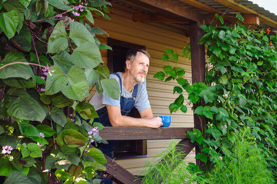 Man in overalls enjoying hot beverage on his rural porch surrounded by lush greenery
