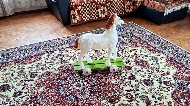 A cute wooden toy horse on colorful wheels stands on a traditional patterned rug in a cozy living room, surrounded by a comfortable sofa, wooden flooring, and natural daylight