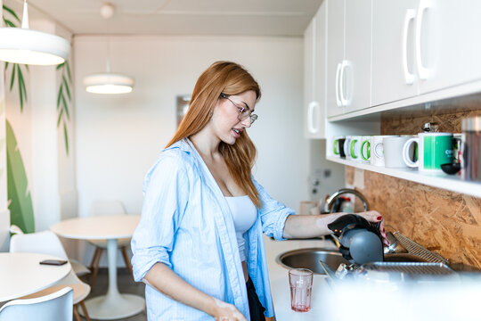 Casual businesswoman making coffee in office kitchen. Young female employee wearing glasses preparing hot drink with capsule machine during break at workplace. 