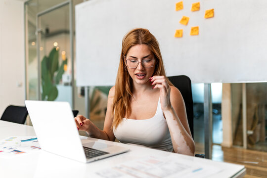 Focused businesswoman with glasses working on laptop in modern office. Concentrated female employee typing and analyzing data at desk during corporate project management. 