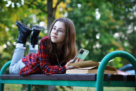 Teen Girl Listening to Music on Smartphone While Reading Outdoors in Autumn Park