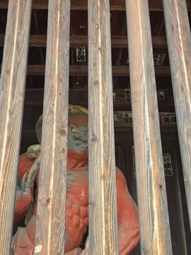 A close-up view of a fierce red Nio guardian statue looking through the wooden slats of a traditional temple gate in Japan.