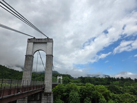 A majestic suspension bridge with tall stone pillars spanning across a lush green gorge under a dynamic cloudy sky in Japan.