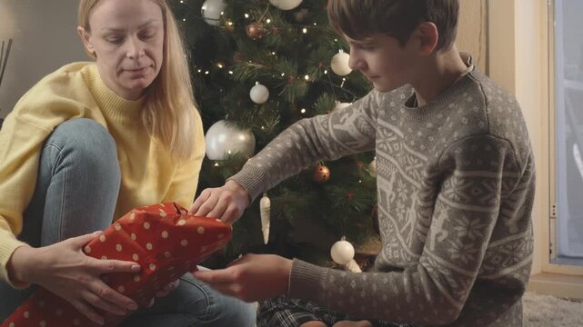 Happy mother spending meaningful time with teenage son while wrapping Christmas present at home, strengthening their parent-child relationship through shared holiday tradition.