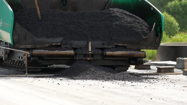 Green asphalt paver spreads hot material onto road. Black asphalt flows steadily from hopper. Machinery moves steadily across construction site. Perfect for road construction projects