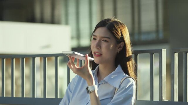 Asian businesswoman leans against a railing and speaks through a smartphone speaker, bathed in soft sunlight in the business district.