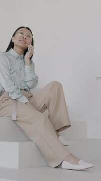 Young Asian woman speaking on mobile phone sitting on stairs in elegant luxury house hall. Cellphone communication and stylish interior concept.