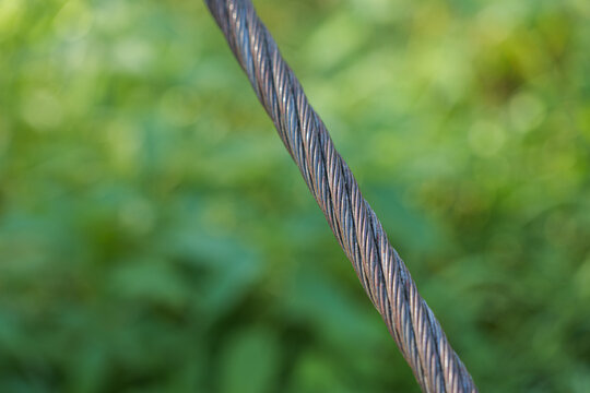 Close-up of heavy steel wire rope on a suspension footbridge, showing twisted metal strands, strength and industrial cable construction against a soft green bokeh background.