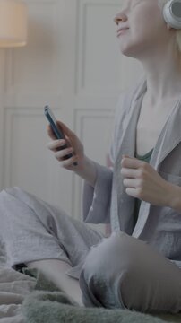 Cheerful albino woman dancing listening to music with headphones holding smartphone sitting in bed in cozy apartment. Modern lifestyle and leisure concept.