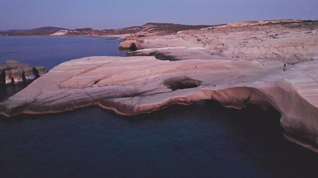 Drone shot of Sarakiniko Beach lunar white rock formations, natural arch, and calm Aegean Sea at twilight in Milos Island, Greece.