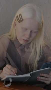 Young albino woman writing in notebook and using smartphone sitting in leather couch in stylish apartment. People and lifestyle concept.