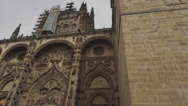 Handheld shot sweeping up the ornate Gothic facade and towering bell tower of Salamanca's iconic Catedral Nueva. Dramatic overcast sky, intricate stonework, dynamic angle.