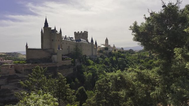 Handheld camera glides past sunlit trees revealing the iconic Alc&aacute;zar de Segovia castle atop a wooded hill, under bright sky. Natural light, dynamic motion, and rich medieval architecture.