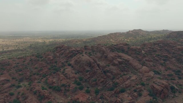 Wide drone panorama over reddish granite hills in the Thar Desert, Sindh, Pakistan, with rounded ridges descending toward hazy sandy plains and sparse scrub.