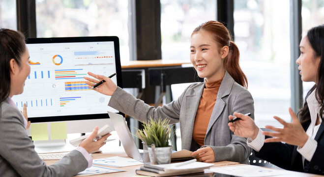 Smiling Asian businesswoman pointing at computer screen presenting charts during team meeting.
