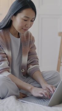 Attractive Asian woman using laptop typing smiling enjoying internet content sitting on bed in luxury house. Computer and modern person concept.