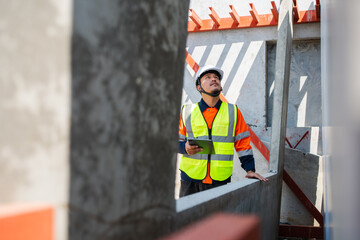 Asian male structural engineer in high visibility jacket holding a tablet while looking up to...