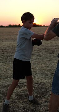 Father teaches son boxing in field at sunset. Boy practices punches with gloves while parent coaches defensive technique. Outdoor practice builds fitness, confidence, discipline, and family