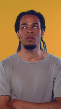 Bored African American man stands with arms crossed against bright yellow background expressing extreme exhaustion by rolling eyes heavily and covering mouth during multiple very deep yawns.