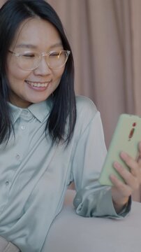 Elegant Asian woman making online video call using smartphone talking and gesturing sitting on couch in luxury apartment. Virtual meeting and distance communication concept.