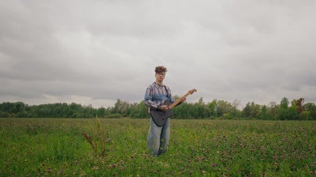 Caucasian young adult playing electric guitar in grassy field as camera zooms in. Suitable for indie music promotion, music streaming campaigns, and audio gear commercials