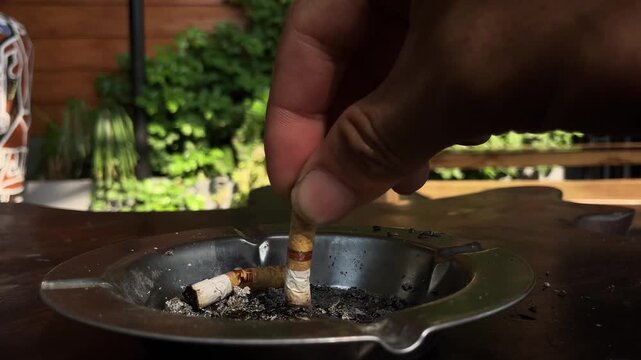 Slow motion close-up video of a person stubbing out and extinguishing a cigarette in a metal ashtray on a wooden table outdoors.