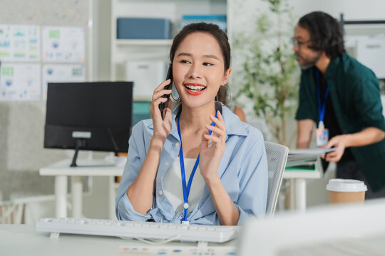 Cheerful Asian businesswoman calling client on smartphone at workplace, communicating friendly corporate advice with professional service satisfaction, confident negotiation, and career enthusiasm.