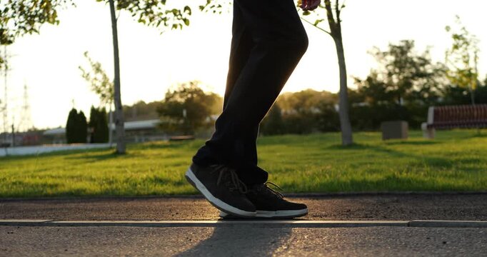 Person walks along park path at sunset in sneakers. Golden sunlight creates calm fitness mood and long shadows. Video supports wellness, exercise, walking, and active lifestyle campaigns.