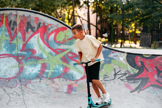 Boy rides scooter in skate park with colorful graffiti on the ramps during the day