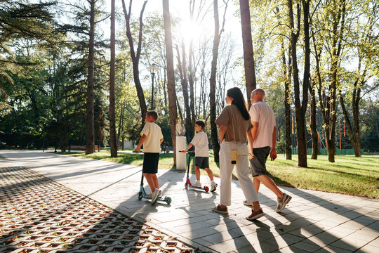 Family walks in park with children riding scooters on a sunny day among trees and pathways