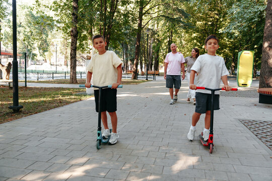 Two boys ride scooters in a park while adults walk nearby on a sunny day