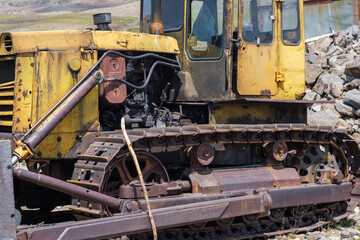 Yellow tracked bulldozer rests on rocky ground, displaying signs of age and use, with a backdrop of industrial materials and machinery © Anat art
