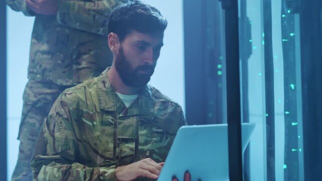 Military member inspects server data meticulously amidst colleagues in secure operational environment