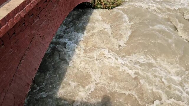 A close-up daytime loop of water flowing steadily through a canal barrage system with red brick piers and mechanical sluice gates. This stock footage is ideal for themes related to water resources, ir