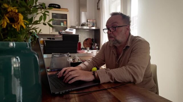 Wide shot of stressed man working online from home while continually squeezing a soft yellow ball, evoking pressure, concentration, relaxation and tactile coping.
