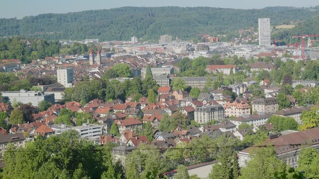 An aerial view showcases a dense cityscape filled with red tiled roofs and interspersed with lush green trees. In the background