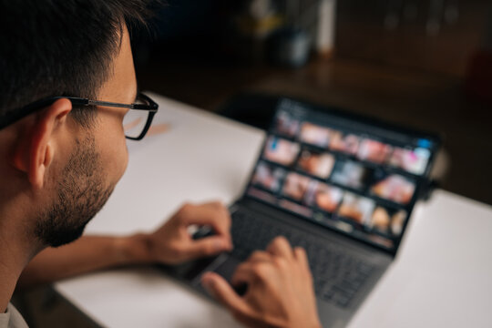 View from shoulder of bearded man in glasses working on laptop at night, screen displaying adult content, potential misuse of technology or lapse in workplace ethics.