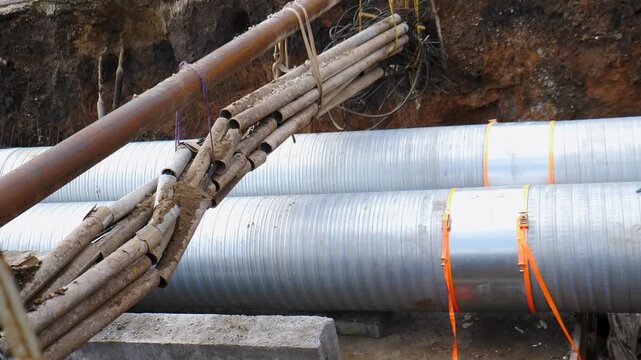 Close-up of large corrugated pipes laid in construction trench. Bundle of wooden planks tied with rope resting on top. Rusty metal pipe above, earth walls, urban utility repair work 4k video