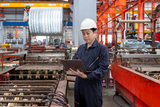A man in a hardhat and mechanic coverall dust protection suit stand in a busy warehouse, manage online document on laptop computer.