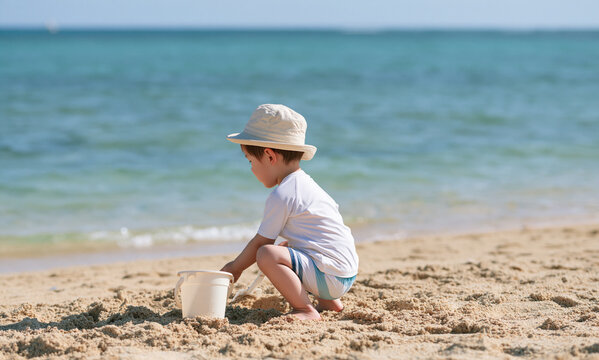  Asian boy wearing a hat, a white rash guard, and swim trunks is crouched on a beach