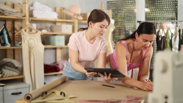 Woman tailor draw dress pattern of product on paper and participate in creating pattern with companion. Craftsman perform paper prototype of product parts, drawing paper pattern for dressmaking. 