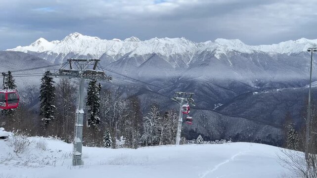 Snow-covered mountains with ski lift in the foreground. Pine trees surround the area. Cloudy sky above, creating a serene winter landscape.