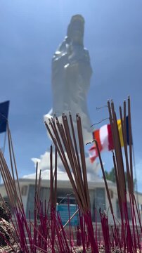 A large statue of a hand is visible in the background, surrounded by colorful flags and incense sticks in the foreground. The scene is set under a clear blue sky.