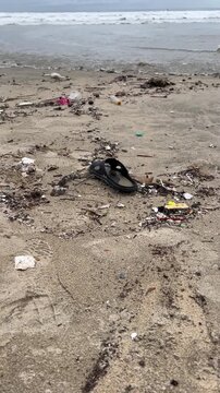 A beach littered with trash, including plastic debris and a single black sandal. The shoreline is visible in the background, highlighting environmental pollution.