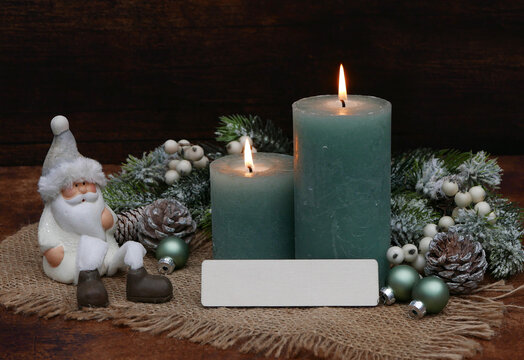A Christmas-themed background with space for text. Two lit green candles, Christmas ornaments, a Santa Claus figure on burlap, and a blank card for writing on.