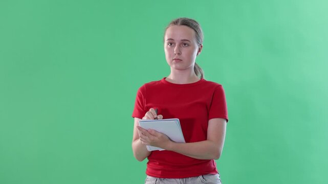 Teen young girl dressed in summer red T-shirt and light blue jeans posing against green backdrop holding documents or writing notes in notebook 