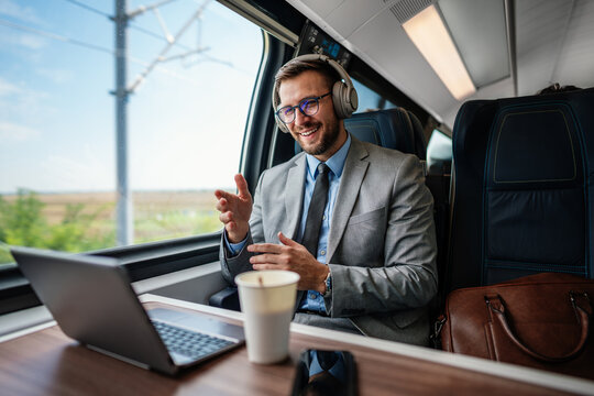Smiling male executive in a suit participating in an online meeting using headphones and computer while traveling in a passenger rail car.