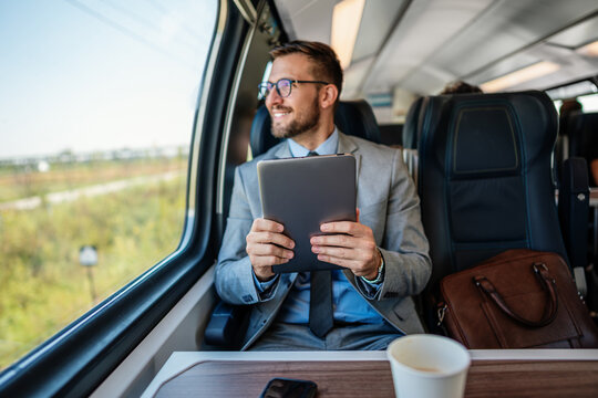 Professional male passenger in business suit analyzing digital data on touchscreen device while sitting by window on modern railway transport vehicle during morning trip.