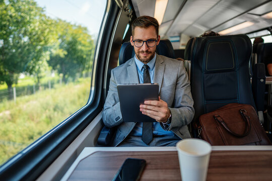Professional male passenger in business suit analyzing digital data on touchscreen device while sitting by window on modern railway transport vehicle during morning trip.
