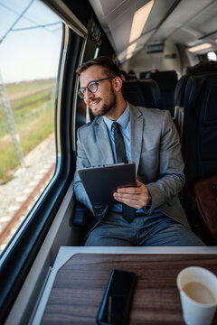 Professional male passenger in business suit analyzing digital data on touchscreen device while sitting by window on modern railway transport vehicle during morning trip.